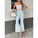 Woman wearing a white top and light blue pants with lace details, standing against a gray wall.